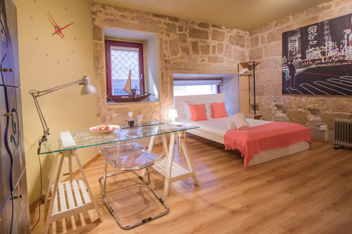 A bedroom with a stone wall, a bed with a red blanket, a desk with a lamp, and a window with a ship model.