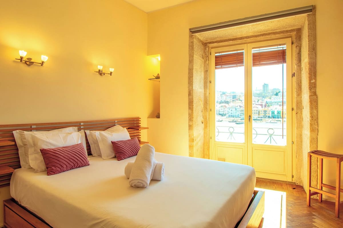 A hotel room with a bed, pillows, a nightstand, a window with a view of the city, and a stone wall.