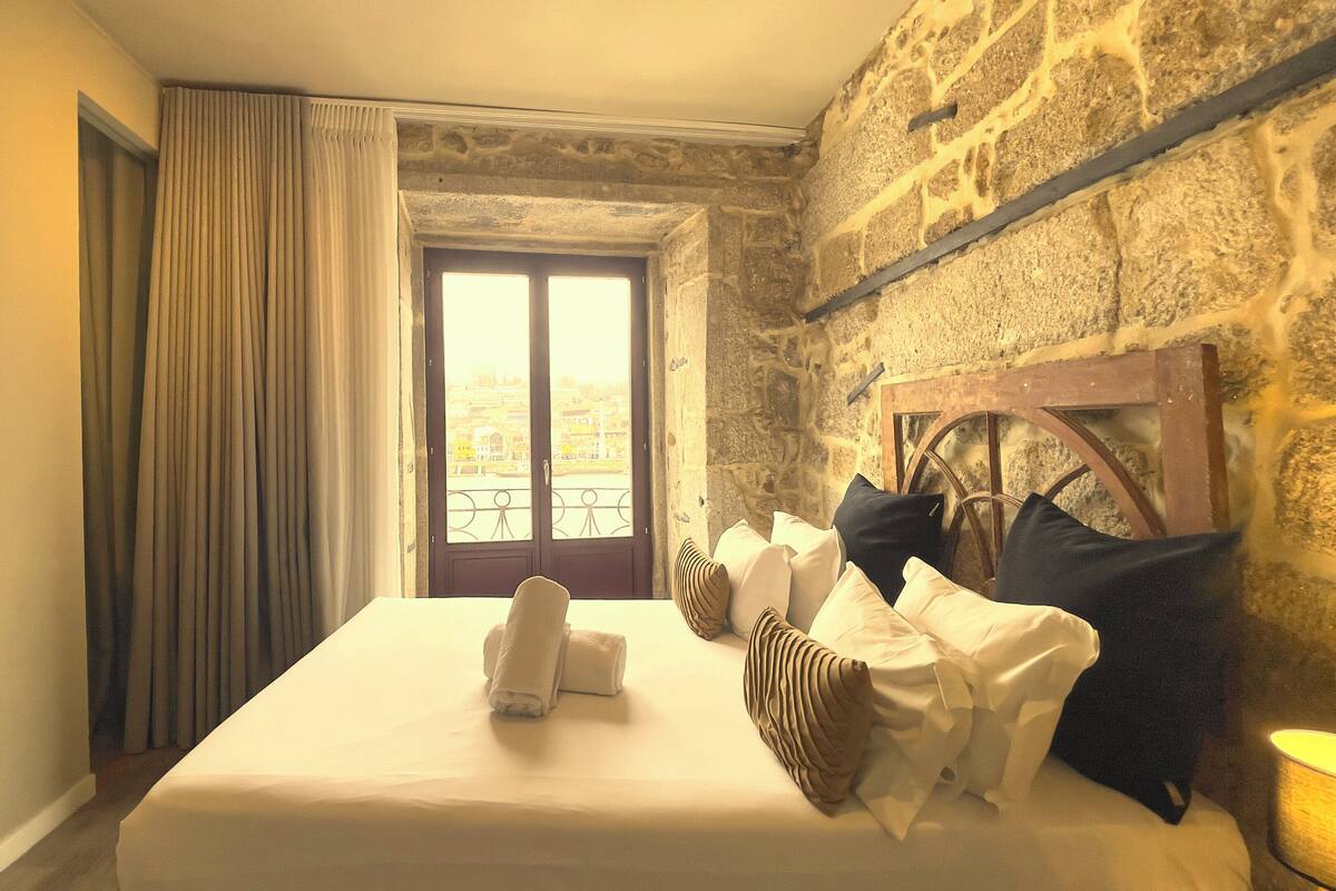 A hotel room with a stone wall, a large bed with white linens, a wooden headboard, and a view through a large window.