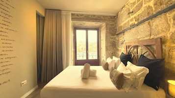 Habitación de hotel con pared de piedra, una cama grande con ropa blanca, cabecera de madera y vistas amplias por una ventana grande.