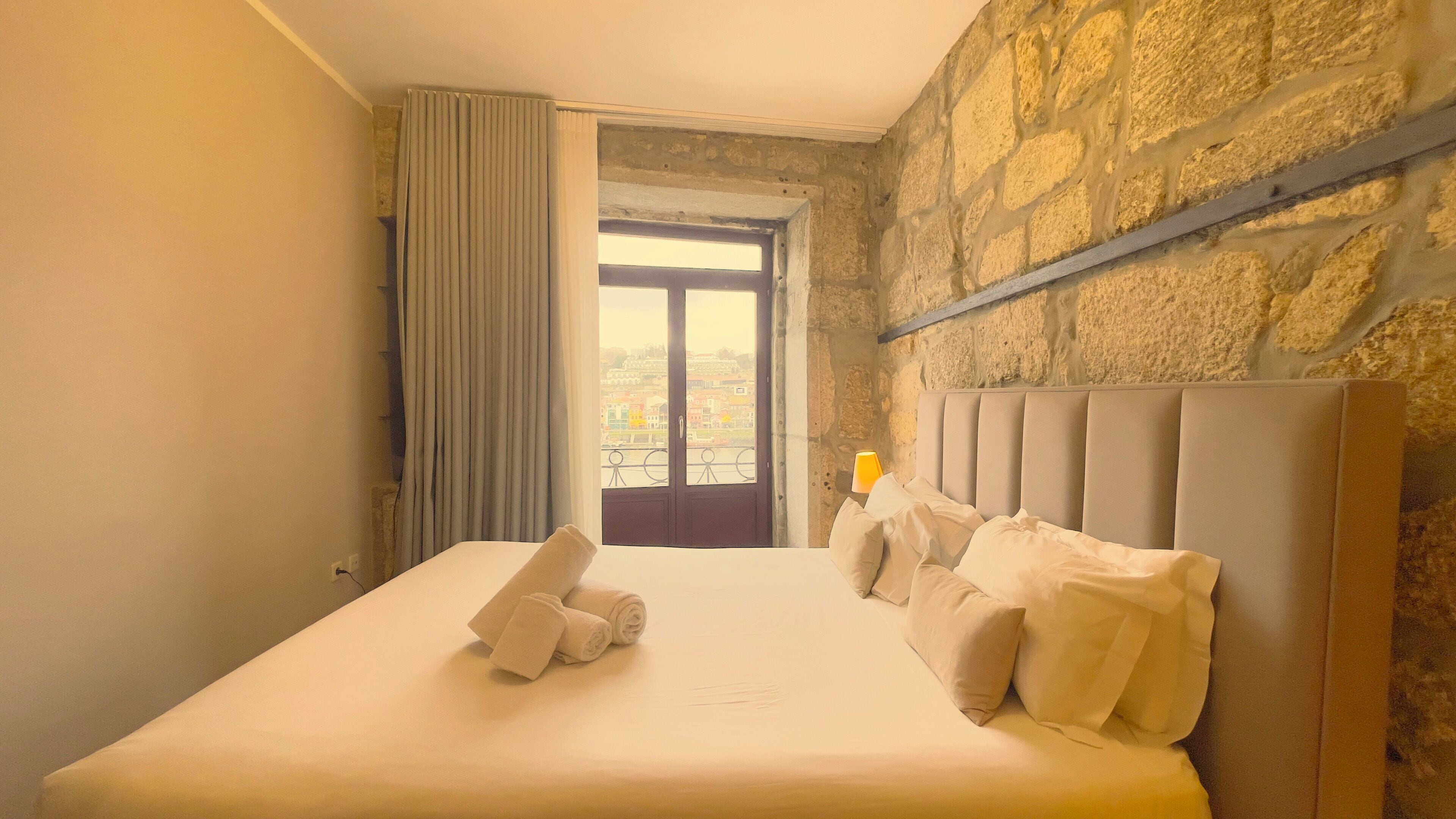 A hotel room with a bed, a window with curtains, a stone wall, and a view of buildings outside.