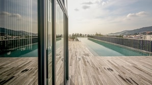 Una terraza de madera con piscina, rodeada por una valla y con vistas a un paisaje urbano y montañas a lo lejos.