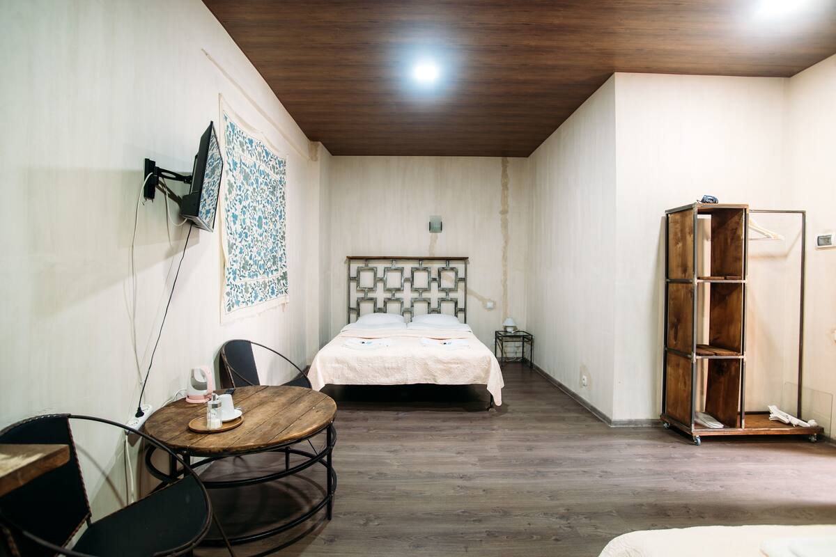 Un dormitorio con suelo de madera, una cama con cabecera estampada, un armario de madera, una mesita con una silla y un televisor montado en la pared.