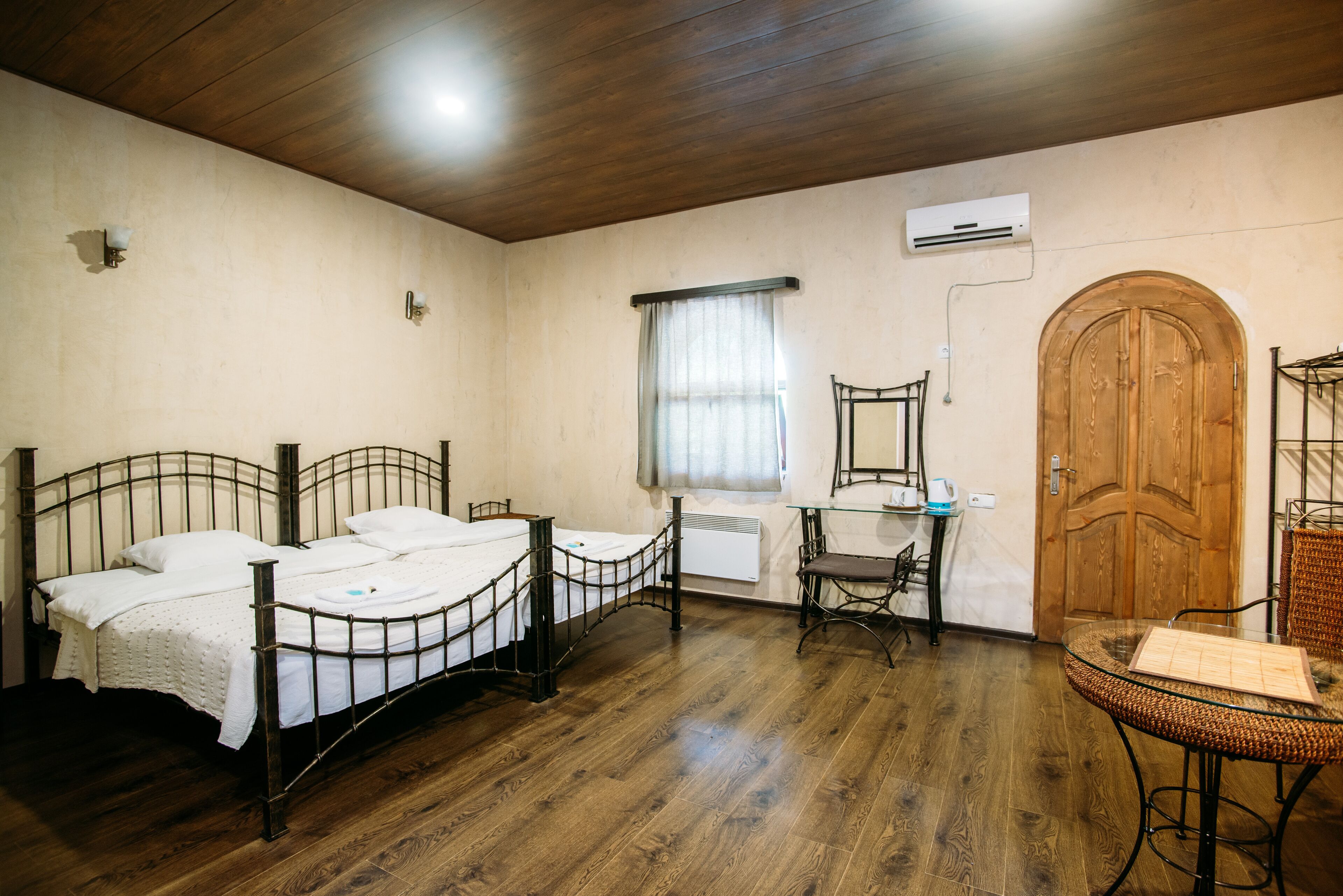 Een kamer met twee bedden met metalen frame, een houten deur, een kleine tafel met stoel en een raam met gordijnen.