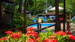 Zona de piscina con sillones de descanso y sombrillas, rodeada de exuberante follaje y flores rojas.