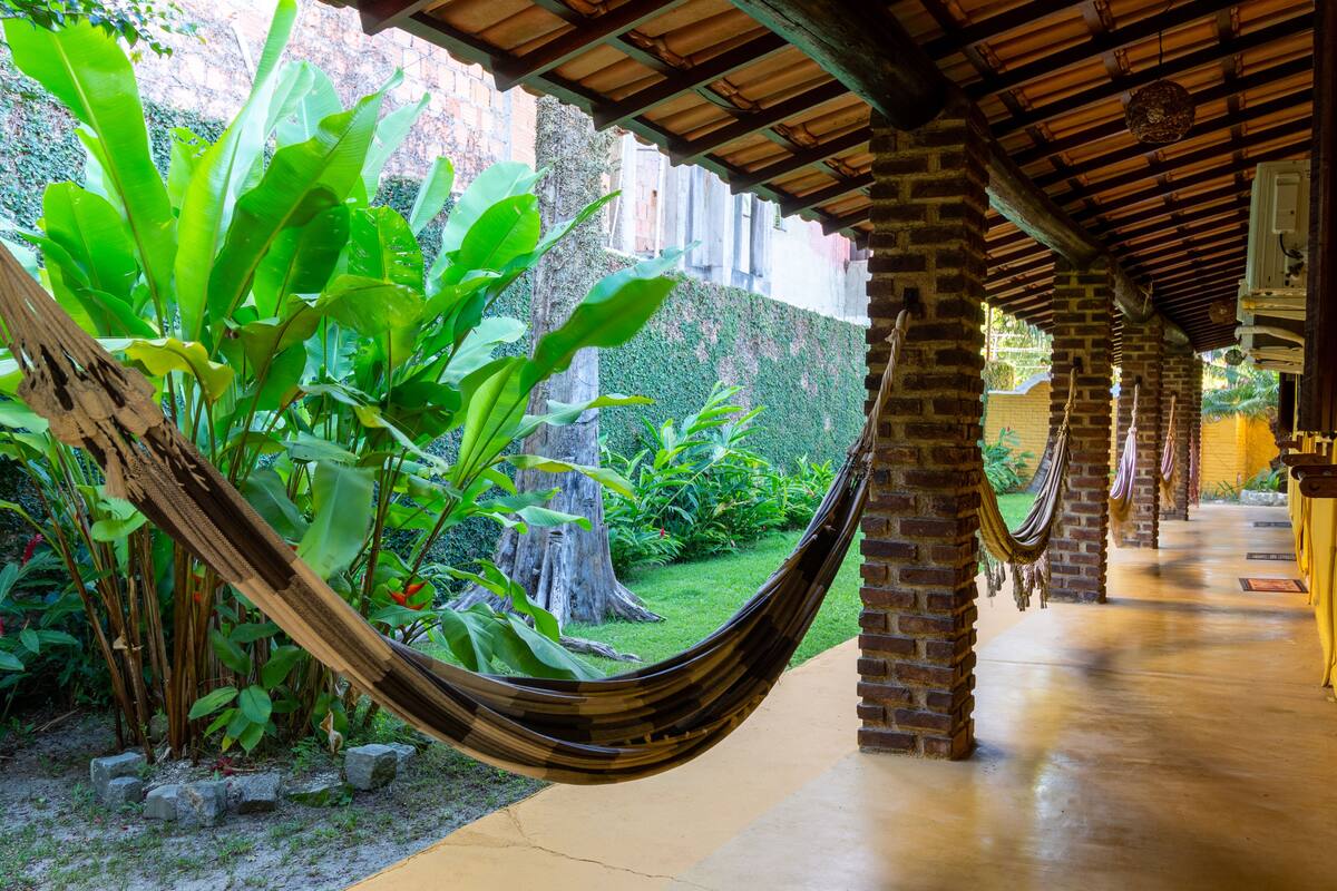 A hammock under a wooden roof, surrounded by lush greenery and brick pillars.