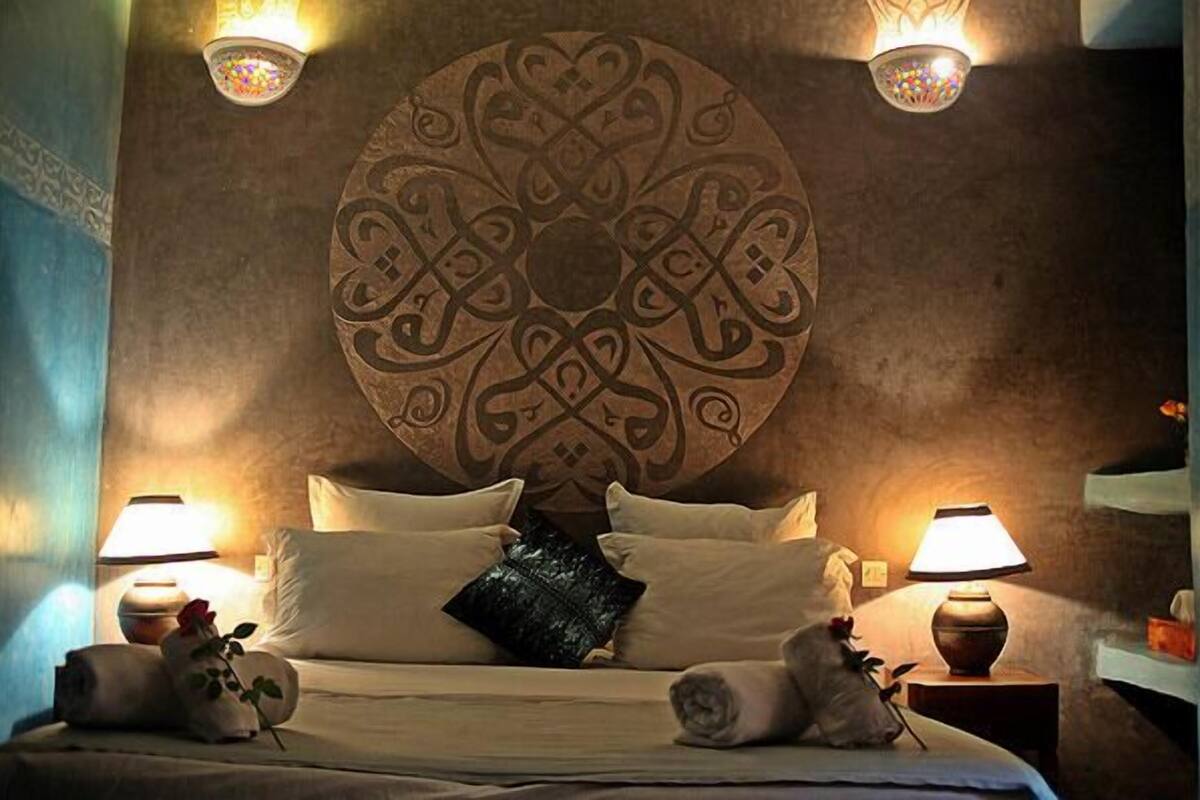 A bed with a circular wall decoration, two bedside lamps, and rolled towels with a rose.