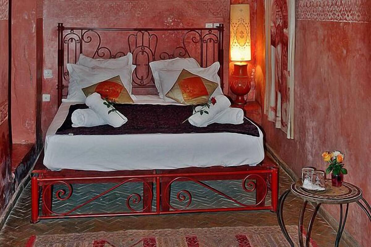 A bed with a metal headboard, white linens, and decorative pillows. A red lamp and a small table with a vase of flowers are visible.