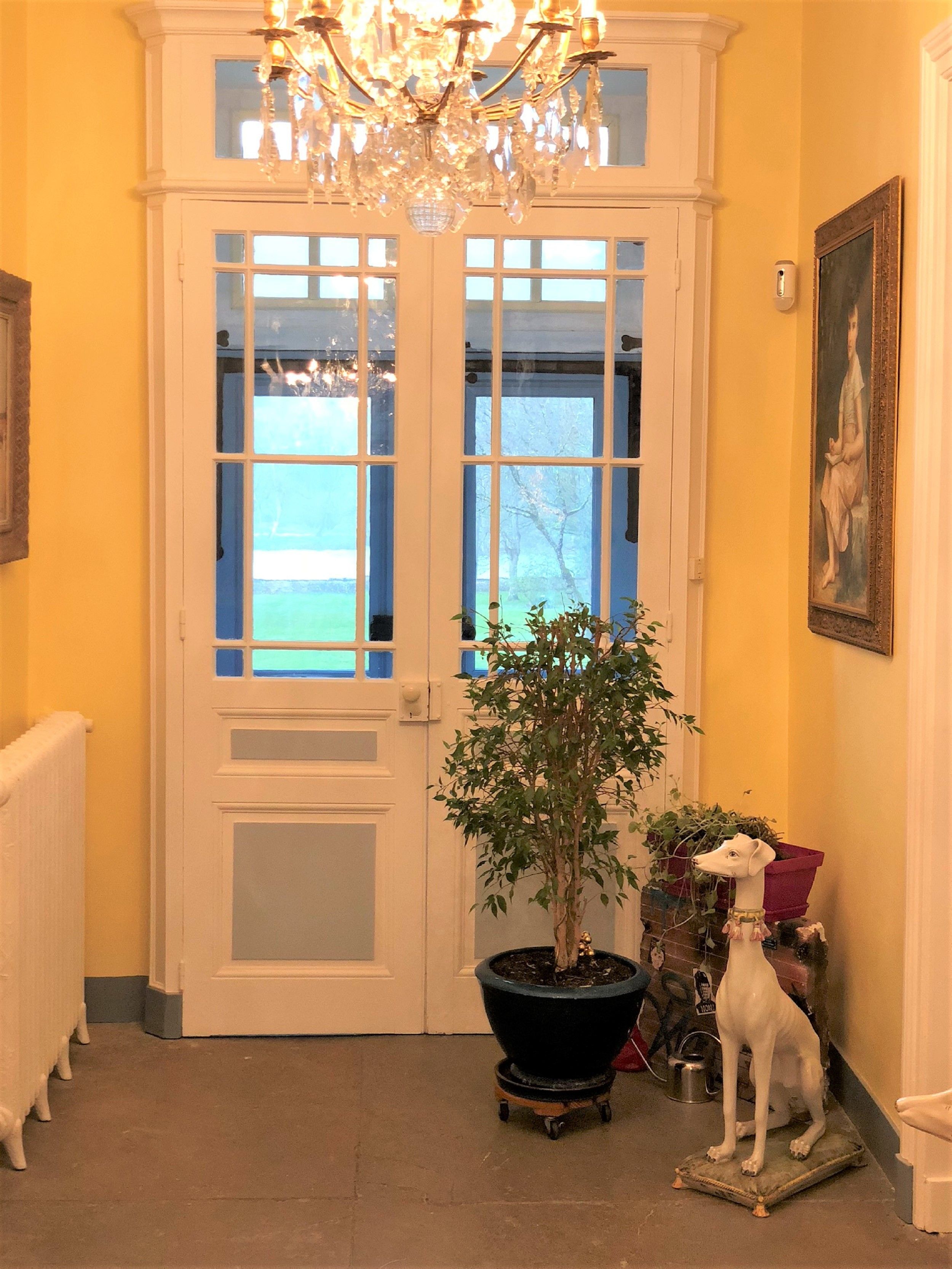 Un pasillo con un puertas dobles, un candelabro, una planta en maceta, una estatua de un perro y un cuadro en la pared.