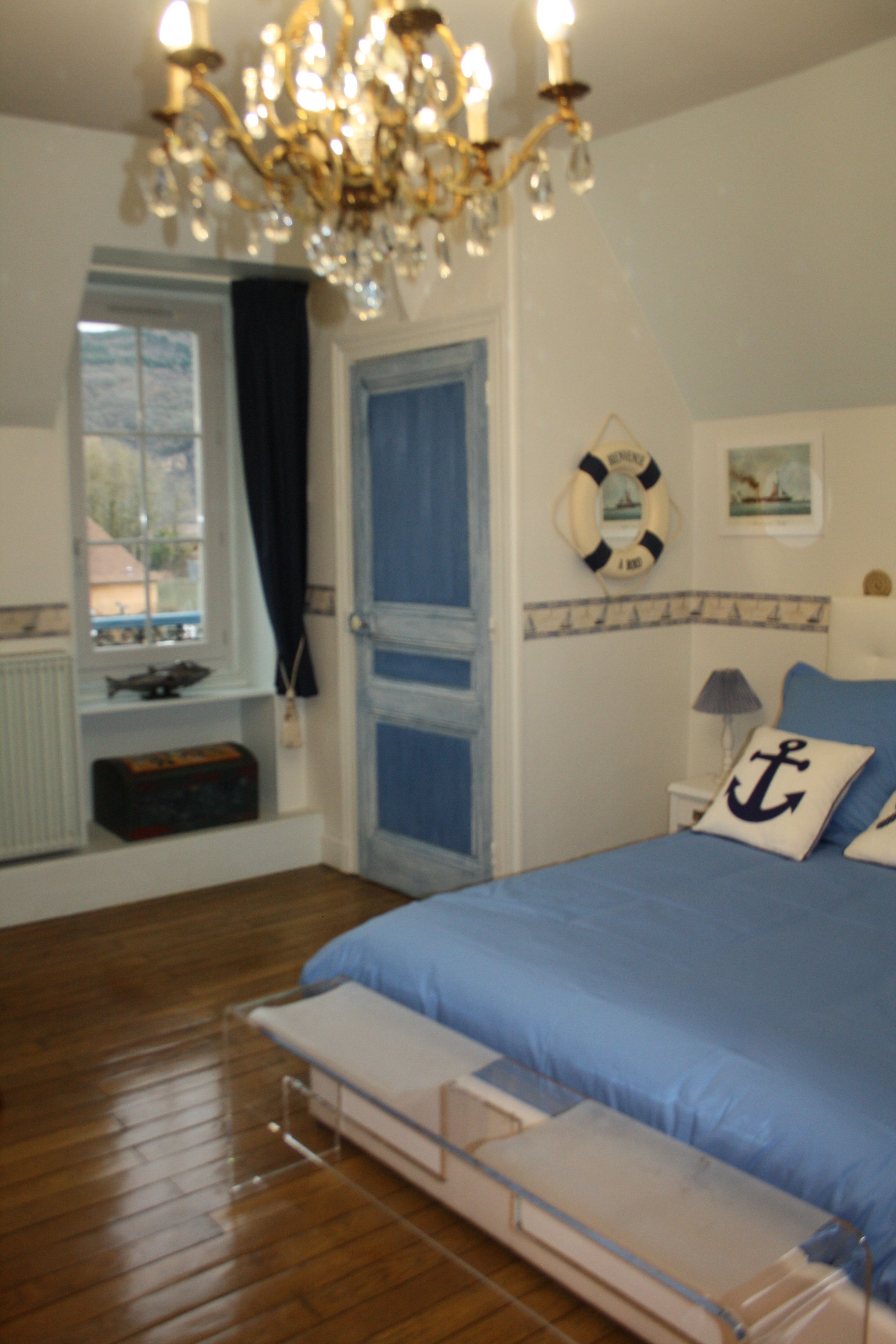 Un dormitorio con cama, candelabro, ventana con cortinas, decoración de salvavidas y una pequeña estantería con objetos.