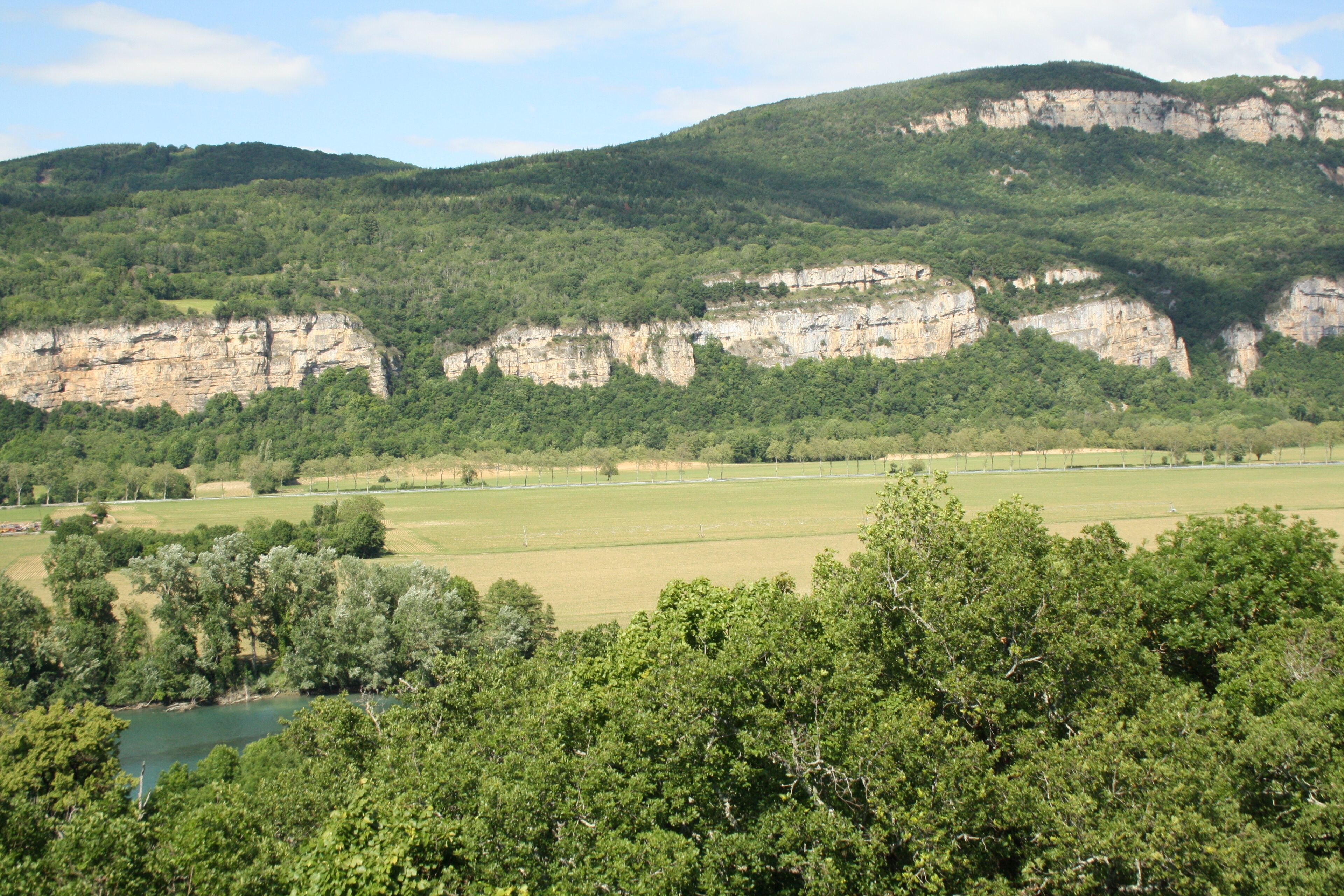 Un paisaje con un río, campos verdes y una colina boscosa con acantilados.