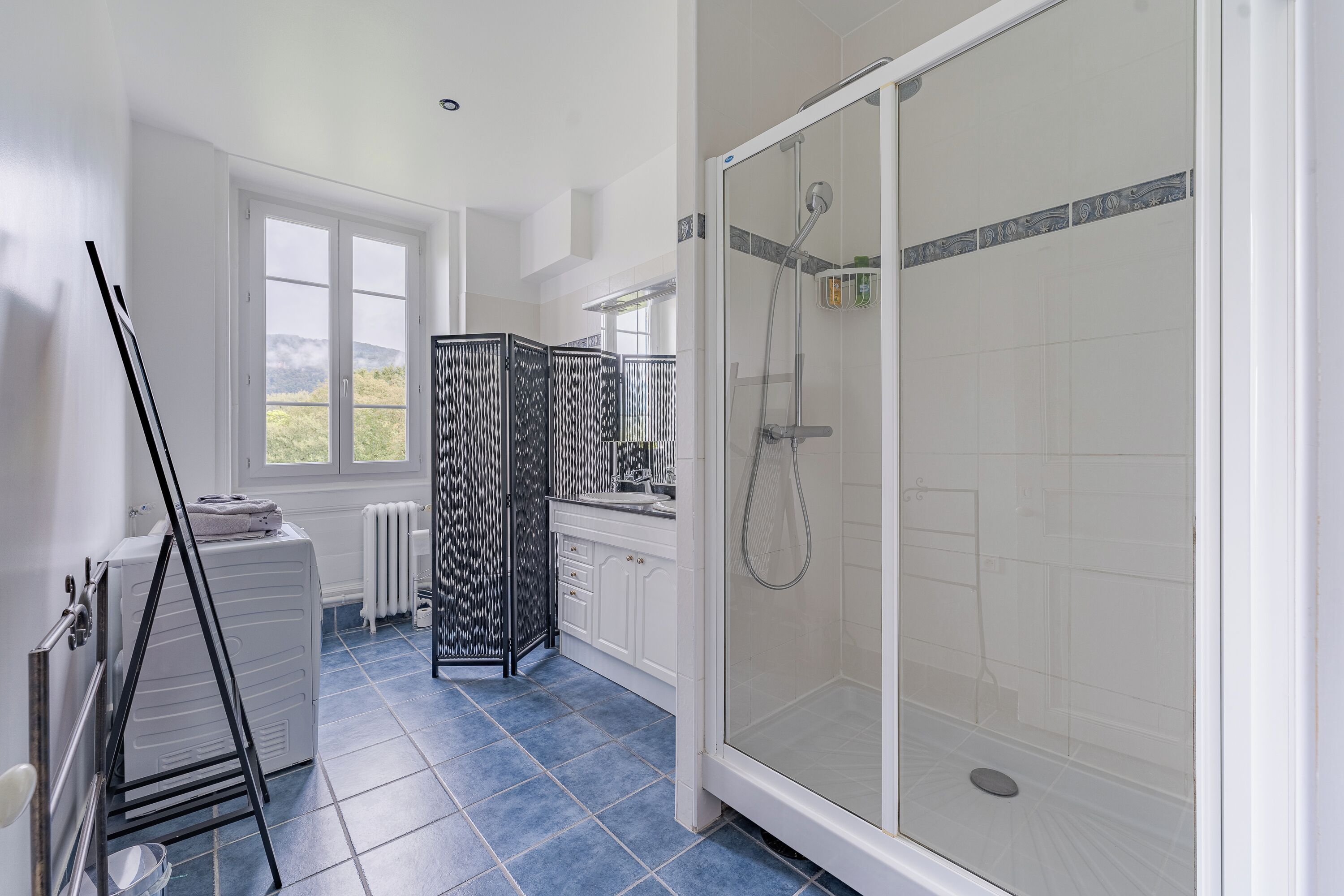 Un baño moderno con ducha de vidrio, muebles blancos y un ventanal grande.