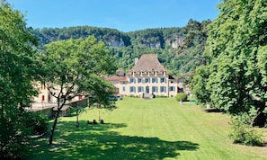 A large, multi-story building with a symmetrical facade, surrounded by lush greenery and a mountainous backdrop.
