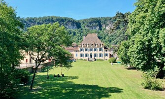 Um edifício grande, com vários andares e fachada simétrica, cercado por vegetação exuberante e um cenário montanhoso.
