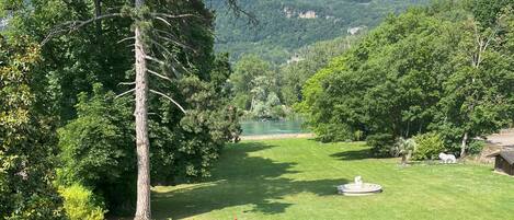 Zona verde bien cuidada con un árbol grande y un arbusto pequeño, un estanque y vistas a un lago y montañas a lo lejos.