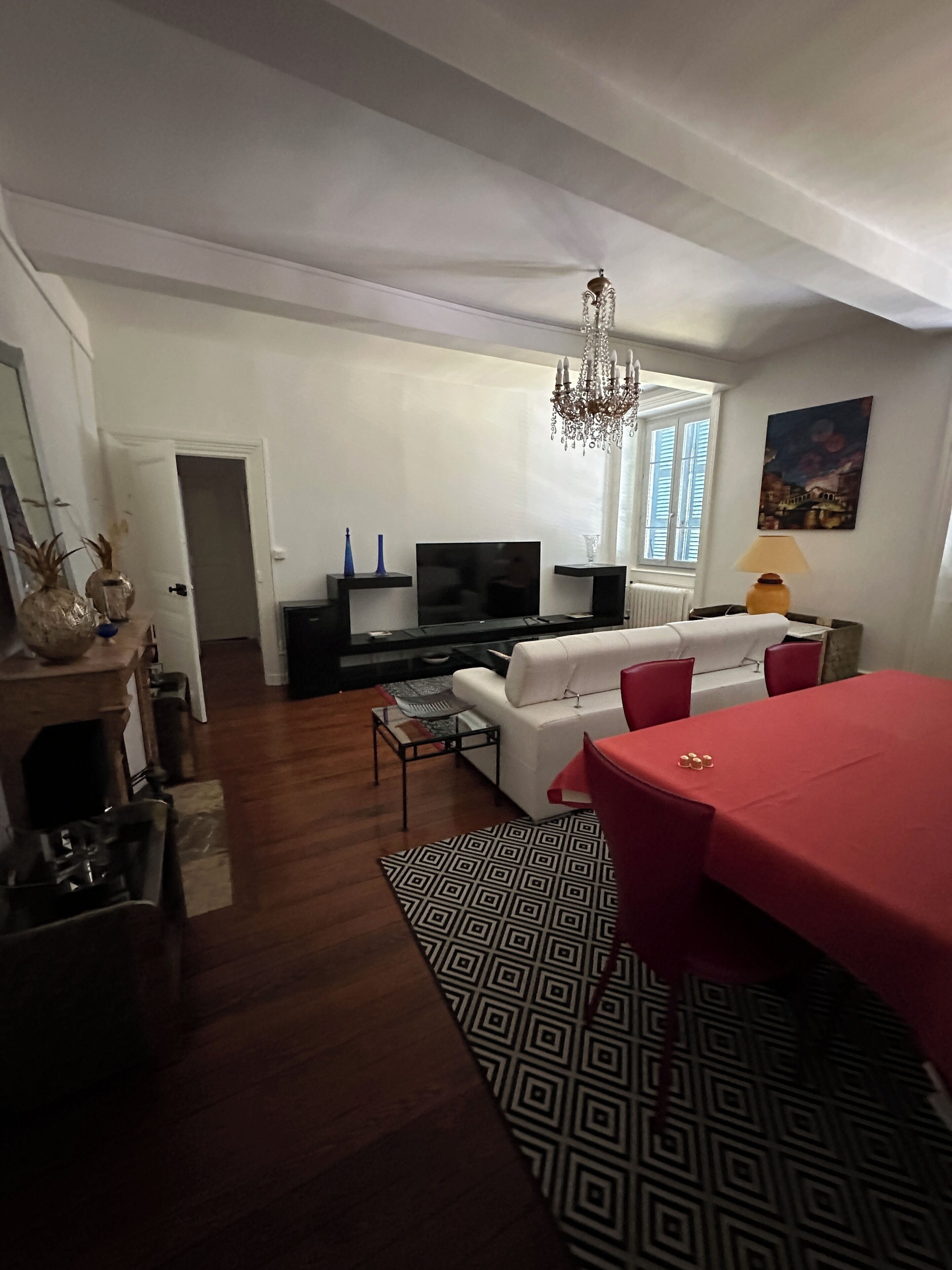 Een woonkamer met een televisie, een witte bank, rode stoelen, een kroonluchter en een eettafel.