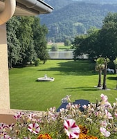 Vista de um gramado bem cuidado com uma fonte, cercado por árvores e um lago ao fundo.