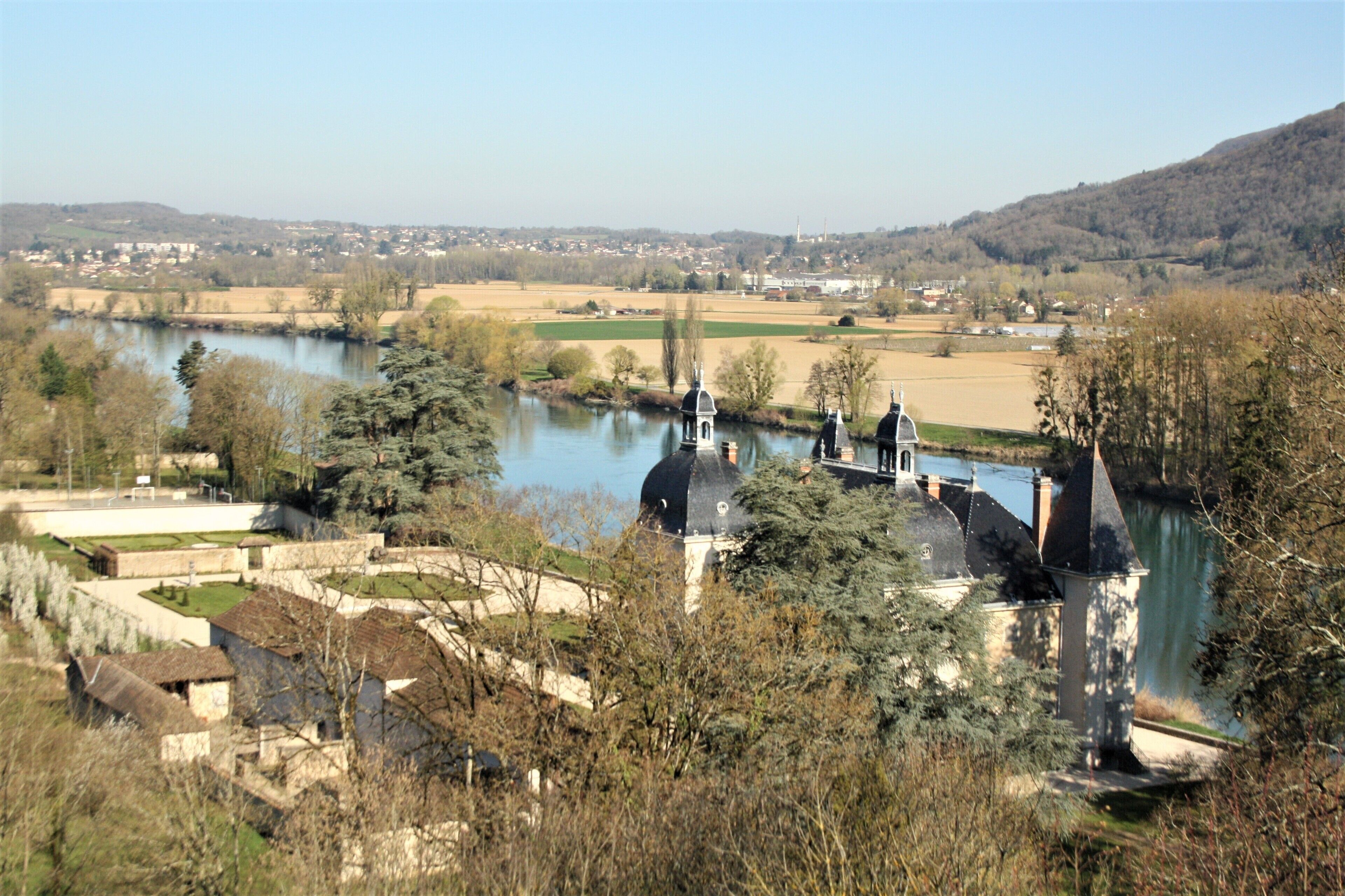 A riverside view with a historic building featuring domed roofs and a tower, surrounded by trees and fields.
