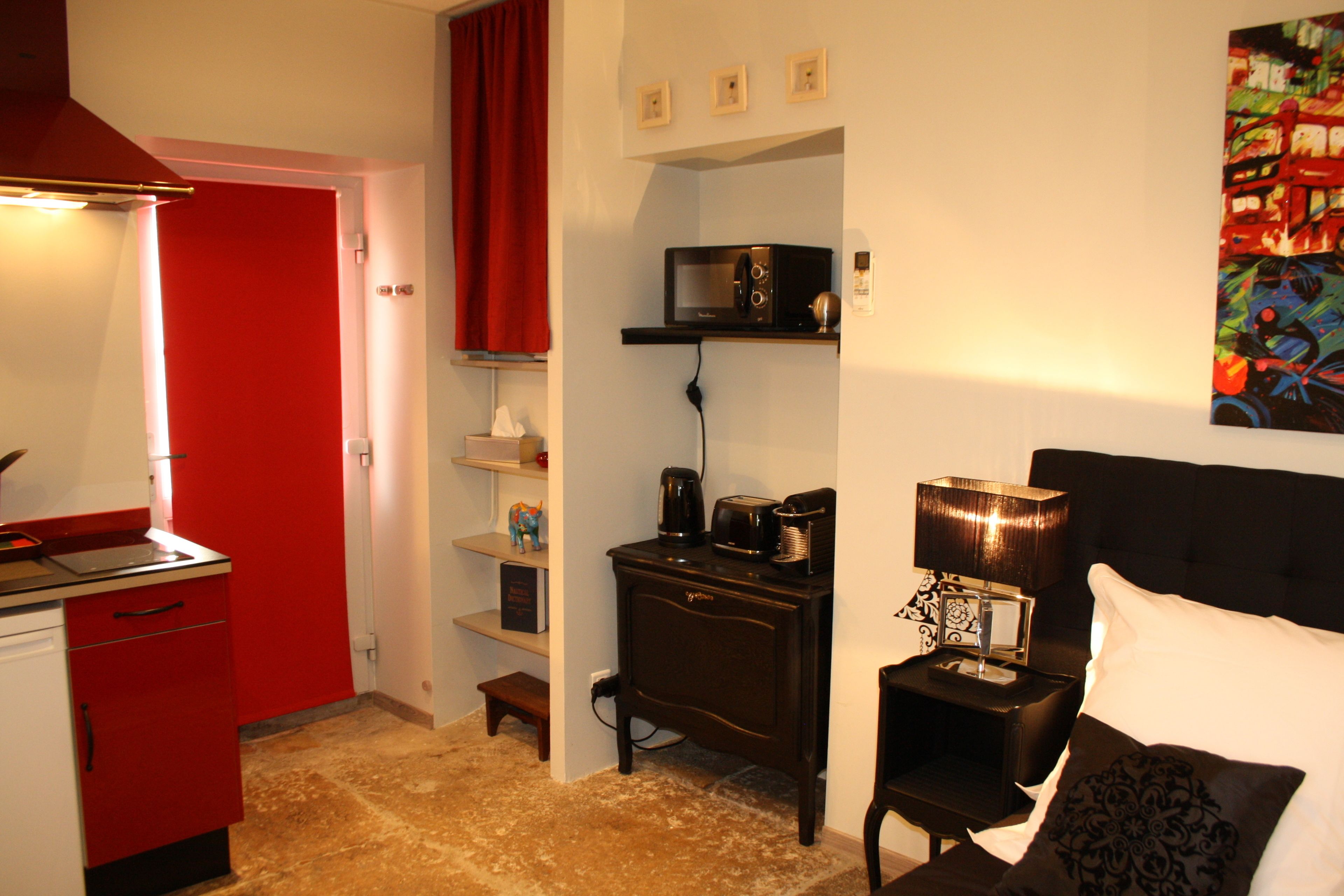 Una habitación de hotel compacta con kitchenette, una cama con cabecera negra, una mesita de noche con lámpara y un cuadro en la pared.