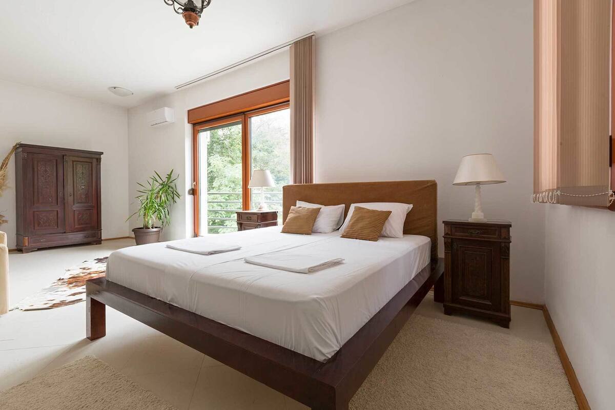 A spacious bedroom with a large bed, a wooden wardrobe, a potted plant, and a view of greenery outside.