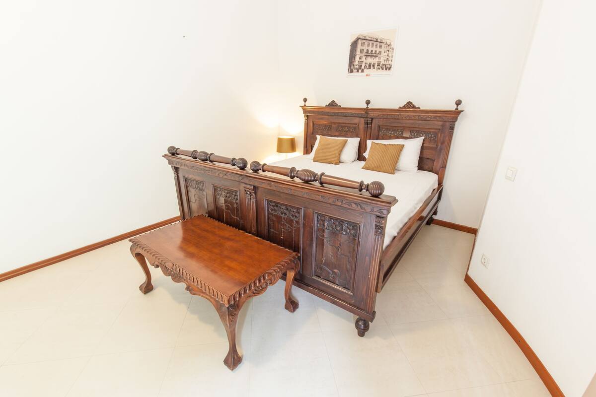 A bedroom with a wooden bed, a wooden nightstand, and a wall-mounted light.
