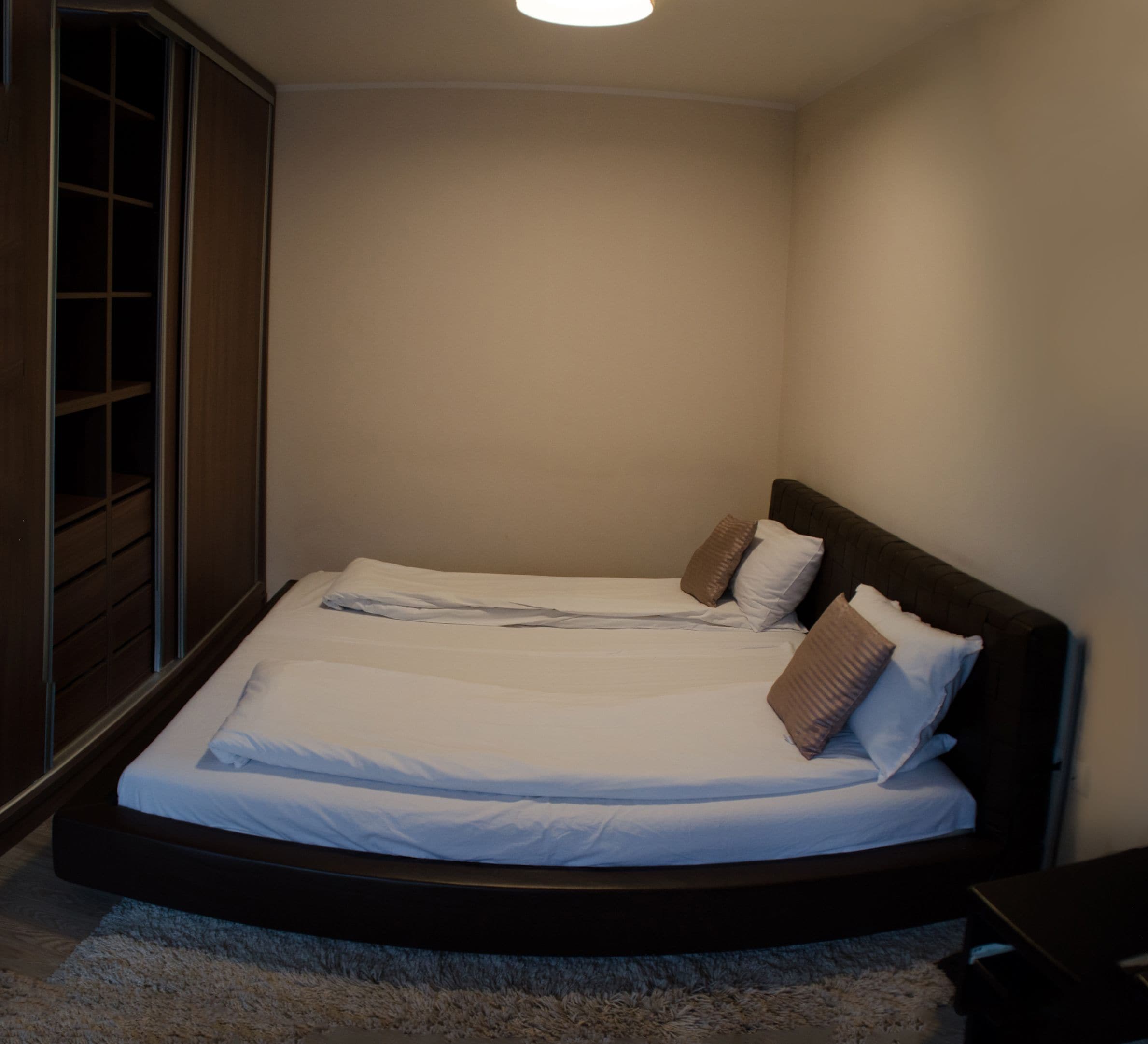 A neatly made bed with white linens and two pillows, a dark headboard, and a wooden wardrobe with a glass door.