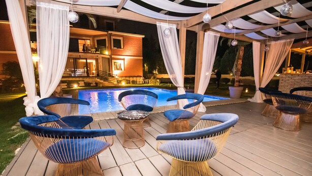An outdoor pool area with blue cushioned chairs and tables, a wooden deck, and a building in the background. - The Belgrade Hills (Belgrade)