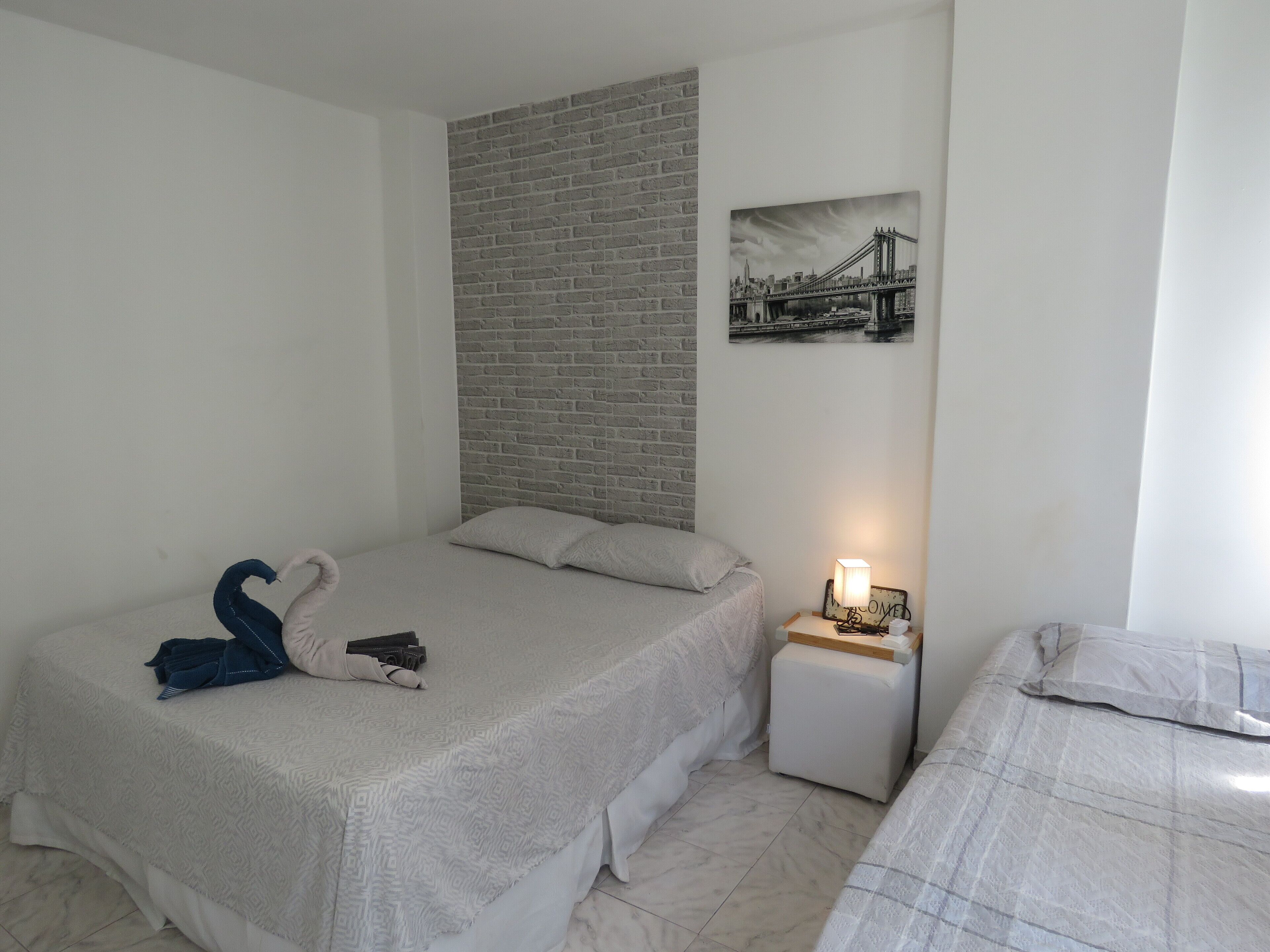 A bedroom with a bed, a nightstand, a lamp, a picture of a bridge, and a brick wall.
