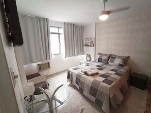 Un dormitorio con cama, escritorio y silla. Hay una ventana con cortinas, un ventilador de techo y un televisor montado en la pared.
