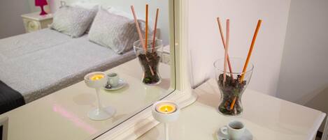 A hotel room with a bed, a bedside table with a candle, a glass of water, and a vase with reed sticks.