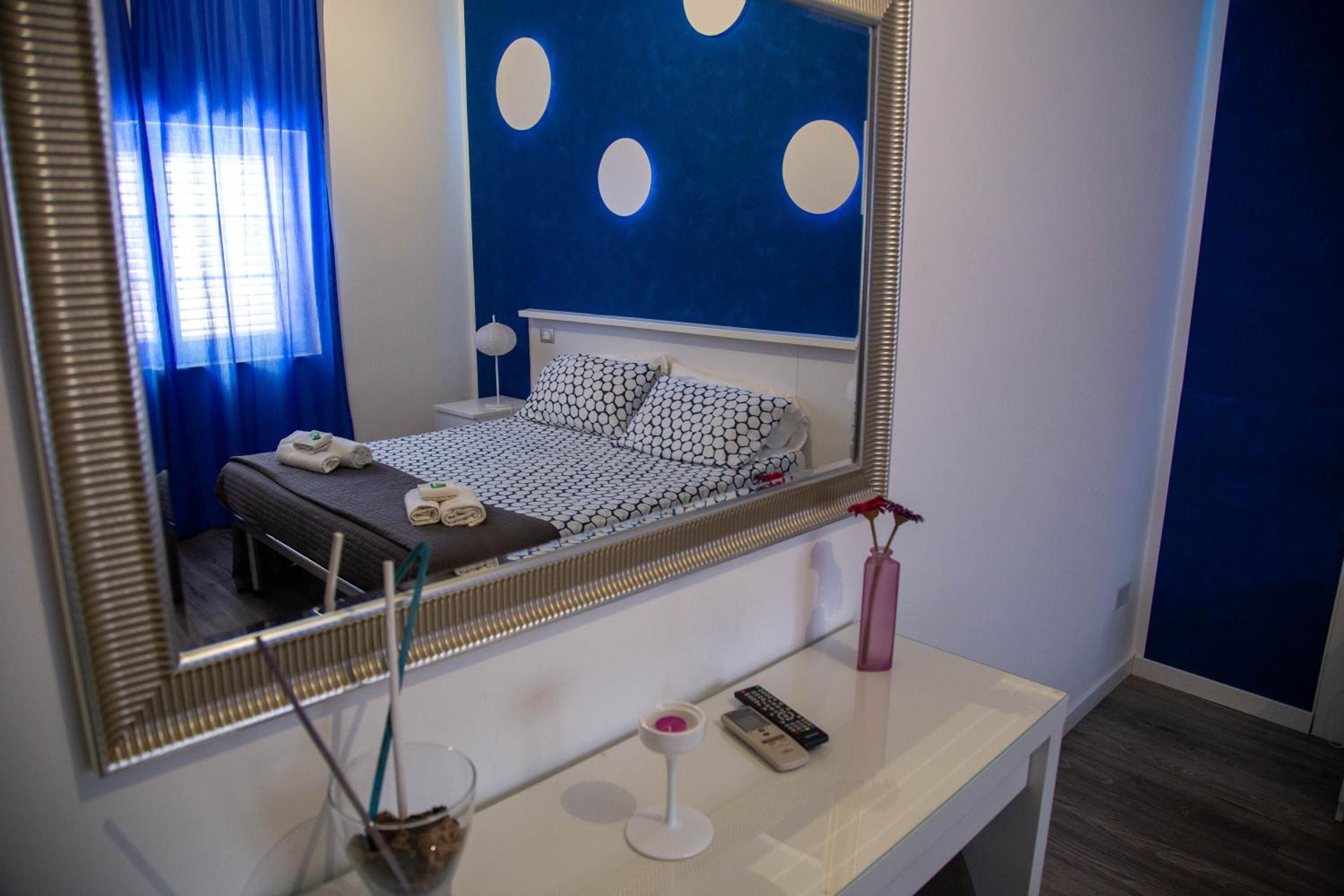Quarto de hotel com um espelho grande, cama com roupa de cama estampada, mesa de cabeceira com um controle remoto, um vaso com uma flor e uma janela com cortinas azuis.