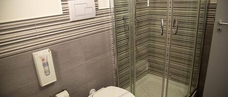 A corner bathroom with a toilet, a glass-enclosed shower, and a towel dispenser.