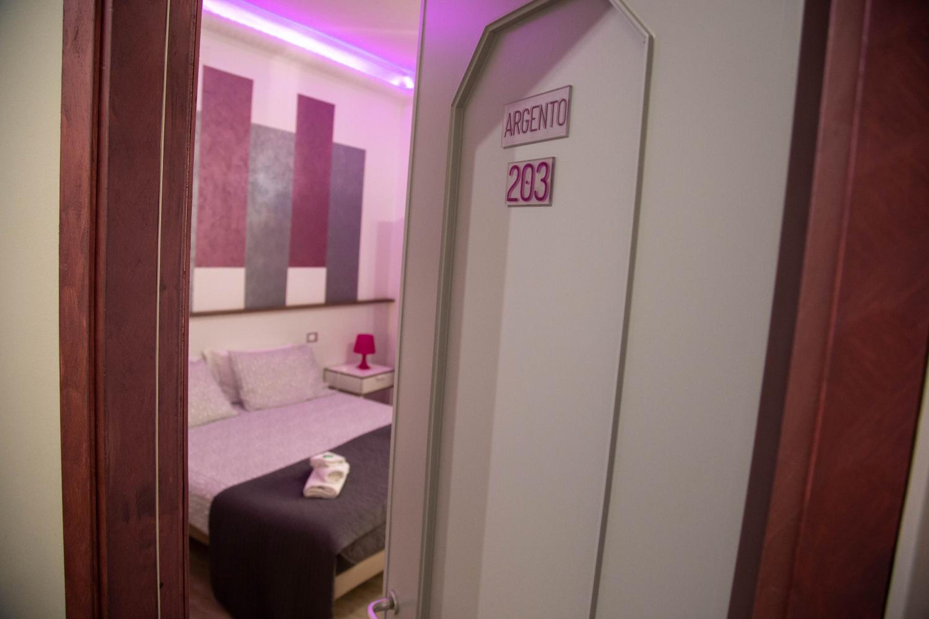 A hotel room with a bed, a nightstand, and a door labeled "ARGENTO 203".