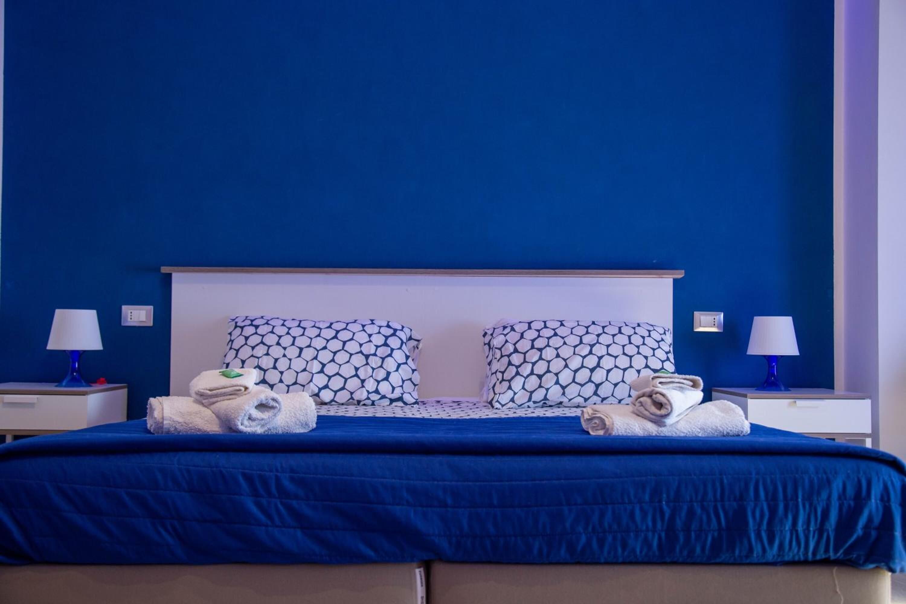 Una cama bien hecha con una colcha azul, dos almohadas estampadas y toallas dobladas en la cabecera.