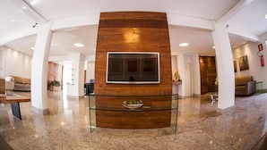 A lobby with a central wooden feature, a glass coffee table, and a flat-screen TV mounted on the wall. - Mundial Parque Hotel (Vicosa)