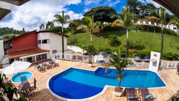 A swimming pool with lounge chairs and umbrellas, surrounded by a fenced patio area with palm trees and a view of a hillside. - Mundial Parque Hotel (Vicosa)
