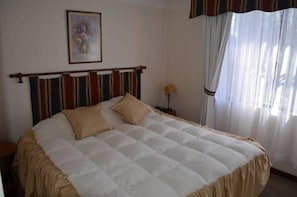 A neatly made bed with a striped headboard, a framed picture on the wall, and sheer curtains on the window. - Apart Hotel Punto Real (Curicó)
