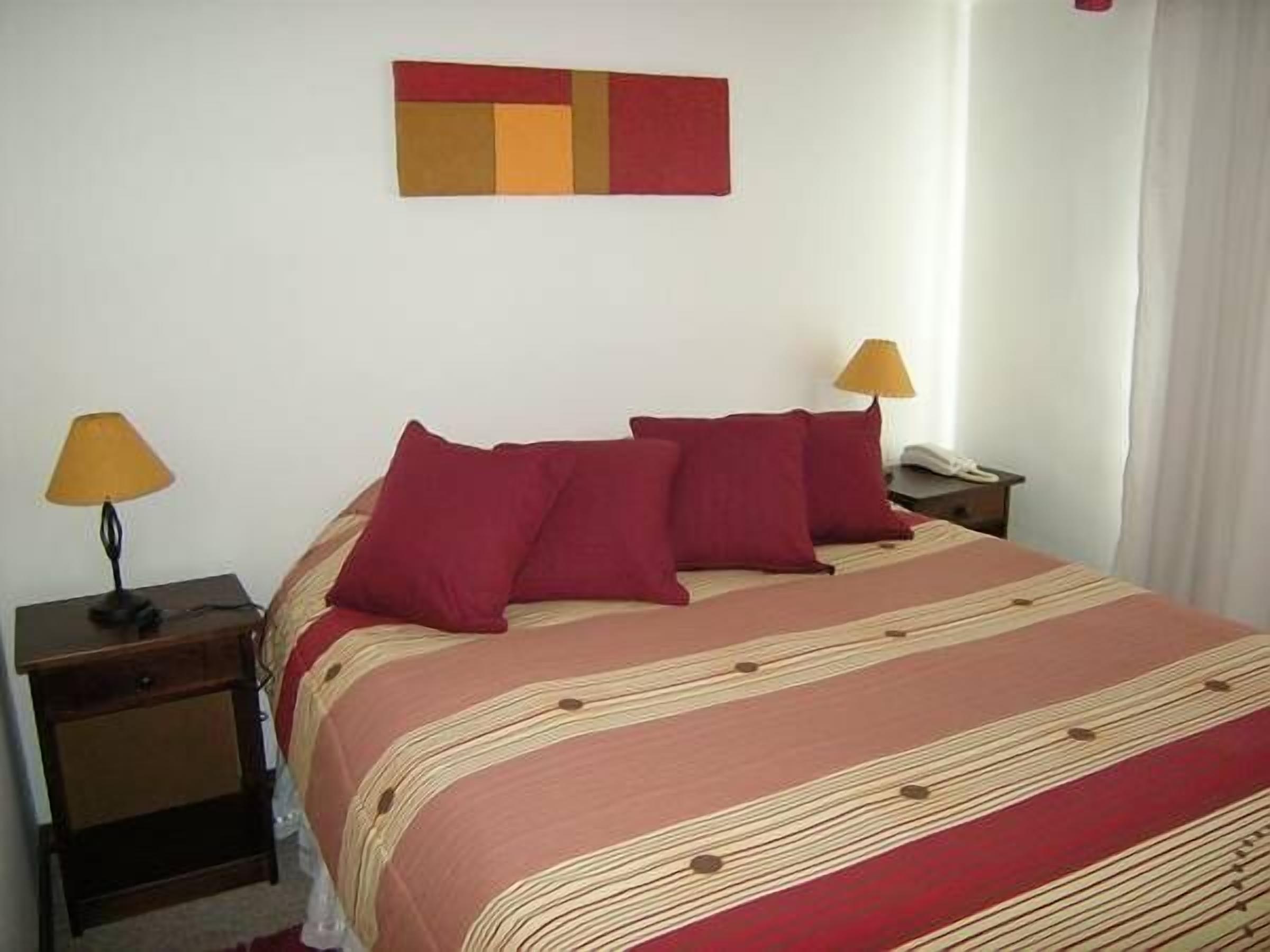 Una cama bien hecha con ropa de cama roja y color beige, dos lámparas de noche y un sencillo cuadro en la pared sobre la cama.