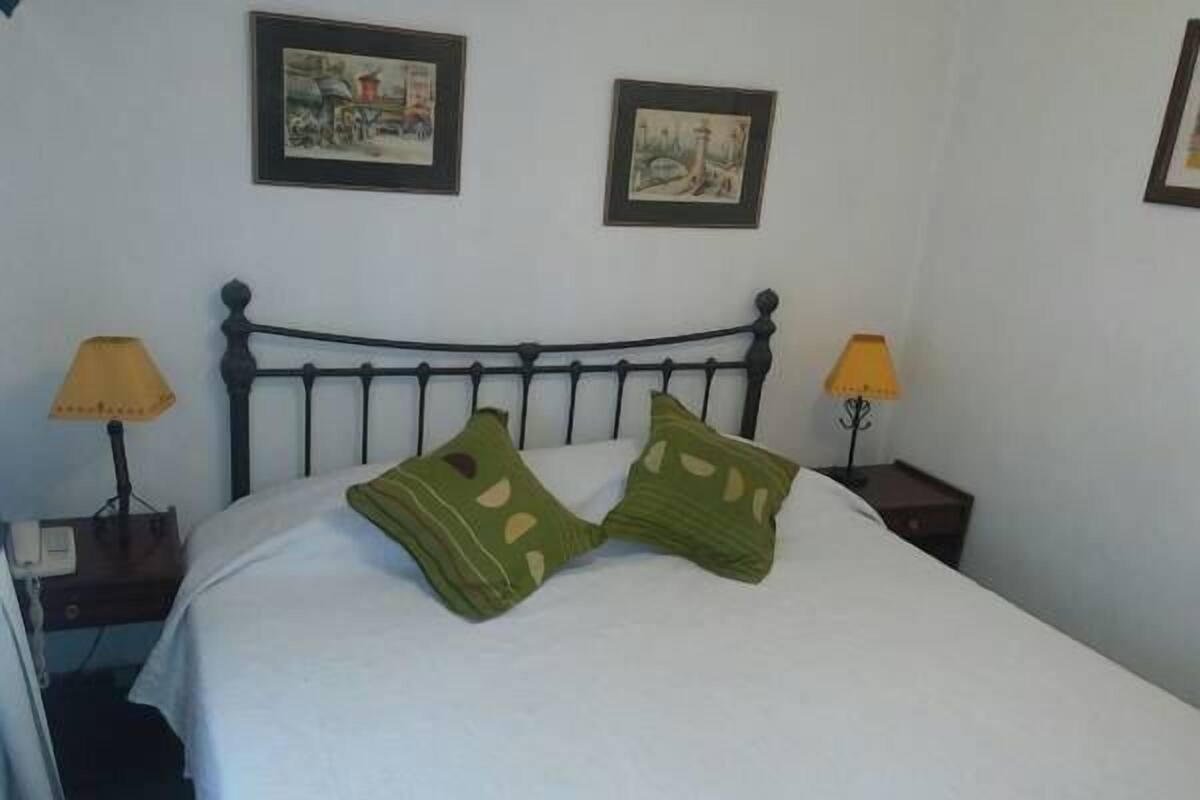 A neatly made bed with two green pillows, a metal headboard, two bedside lamps, and two framed pictures on the wall.