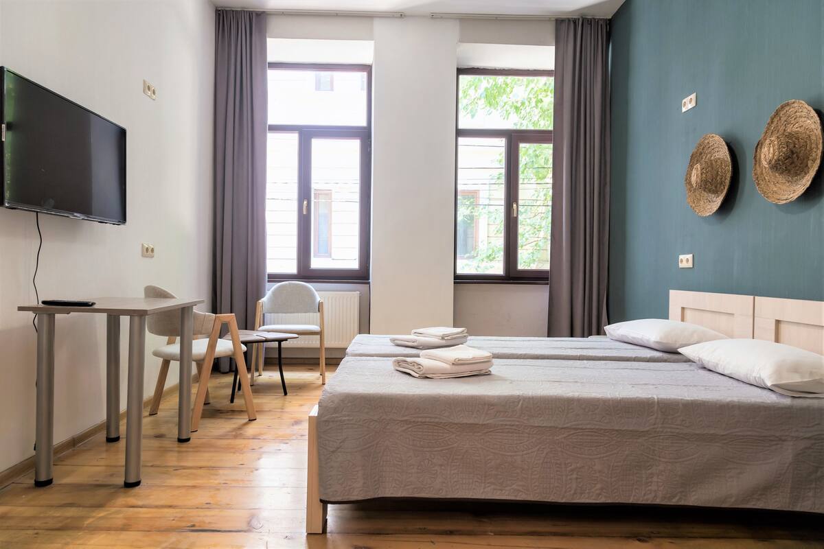 A hotel room with a bed, a desk, a chair, a TV, and two windows with curtains.