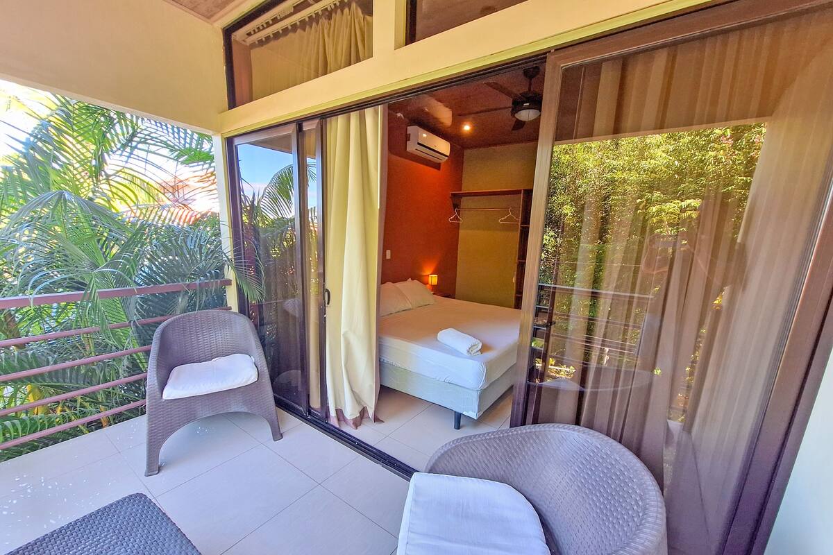 Un balcon avec des fauteuils en osier, un lit avec un matelas et un oreiller blancs, et une vue sur la verdure.