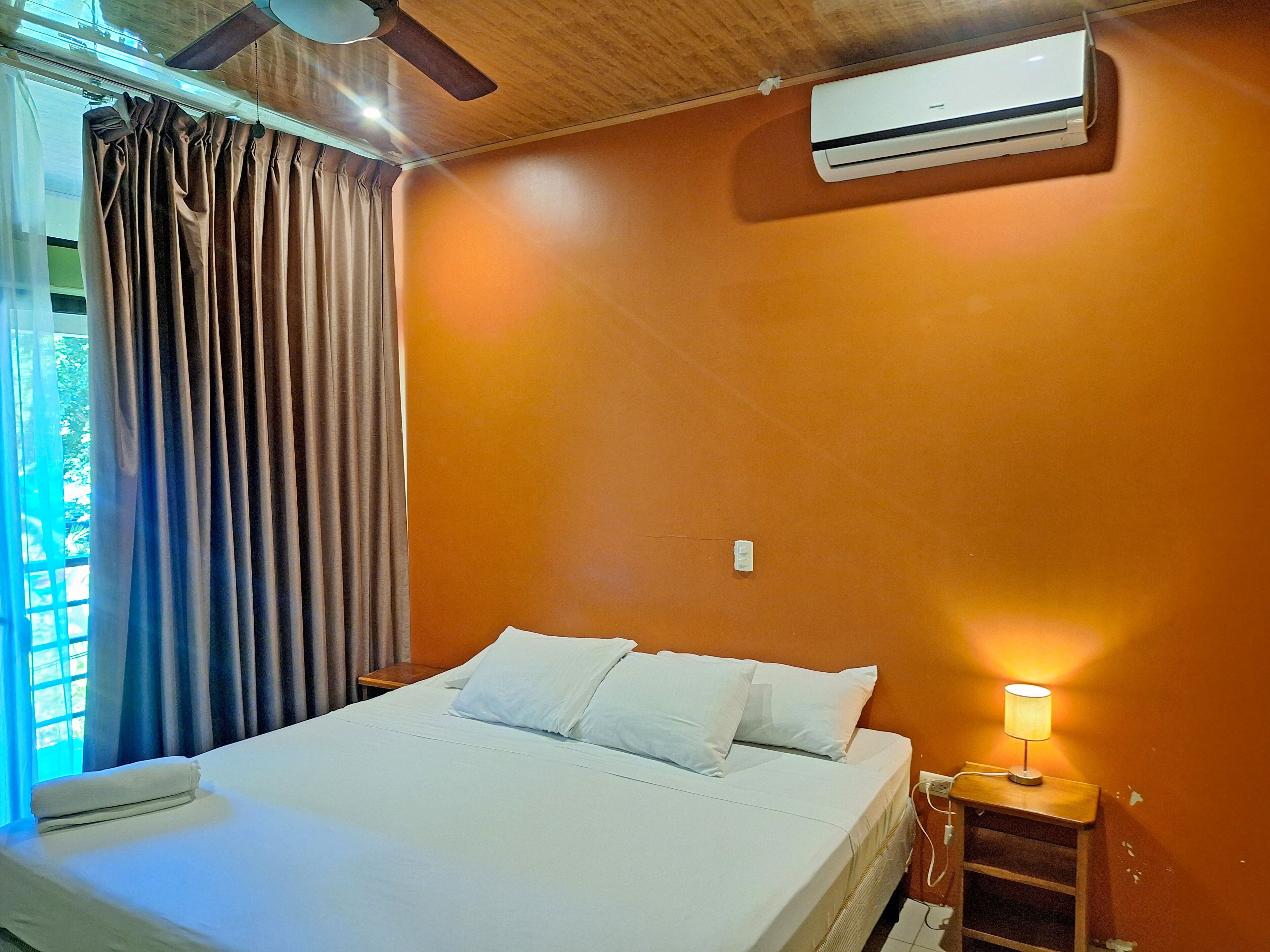 A hotel room with a bed, an air conditioner, a nightstand with a lamp, and a window with curtains.