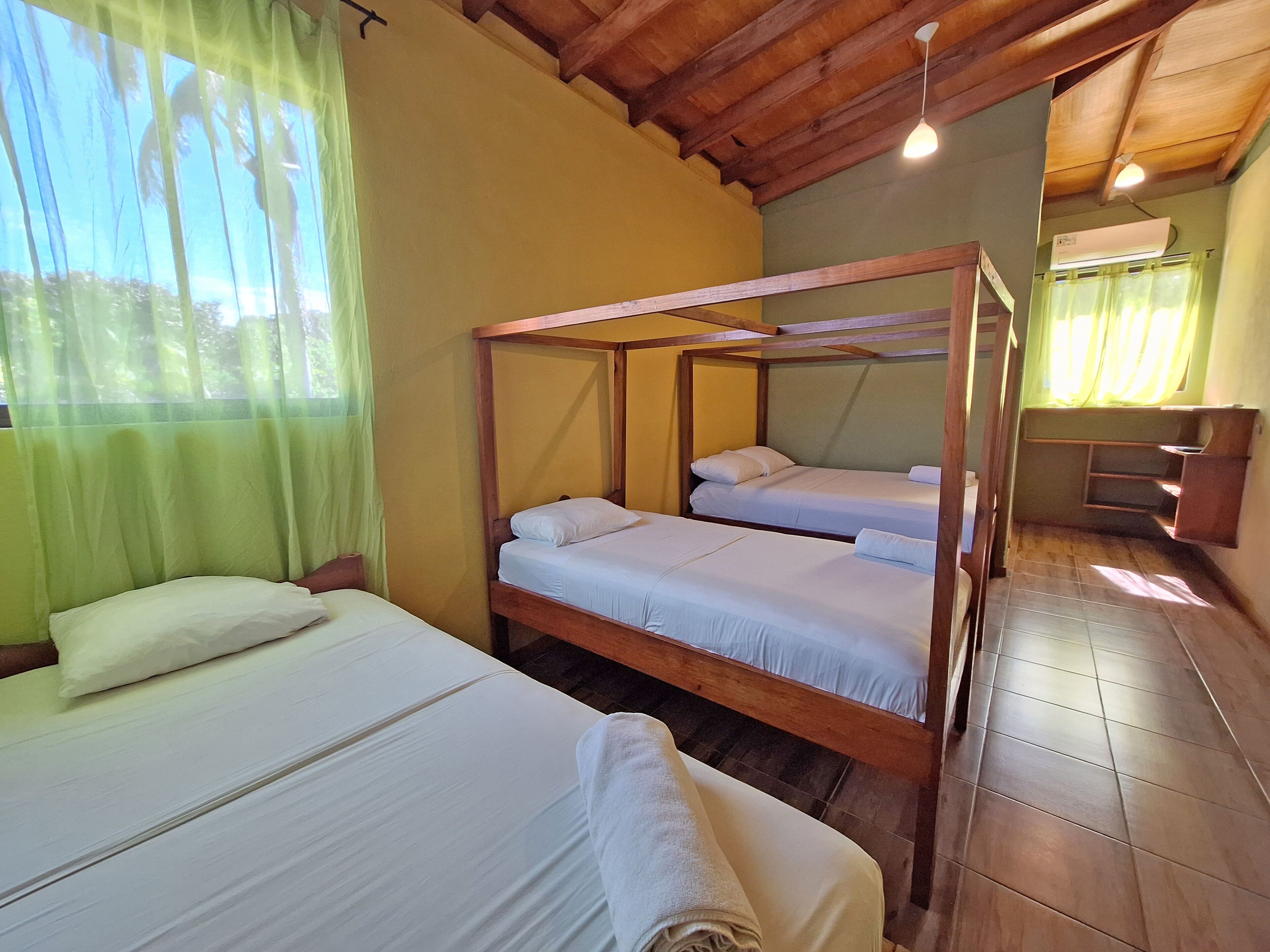 A room with two single beds and a bunk bed, wooden beams on the ceiling, a small desk, and a window with sheer curtains.