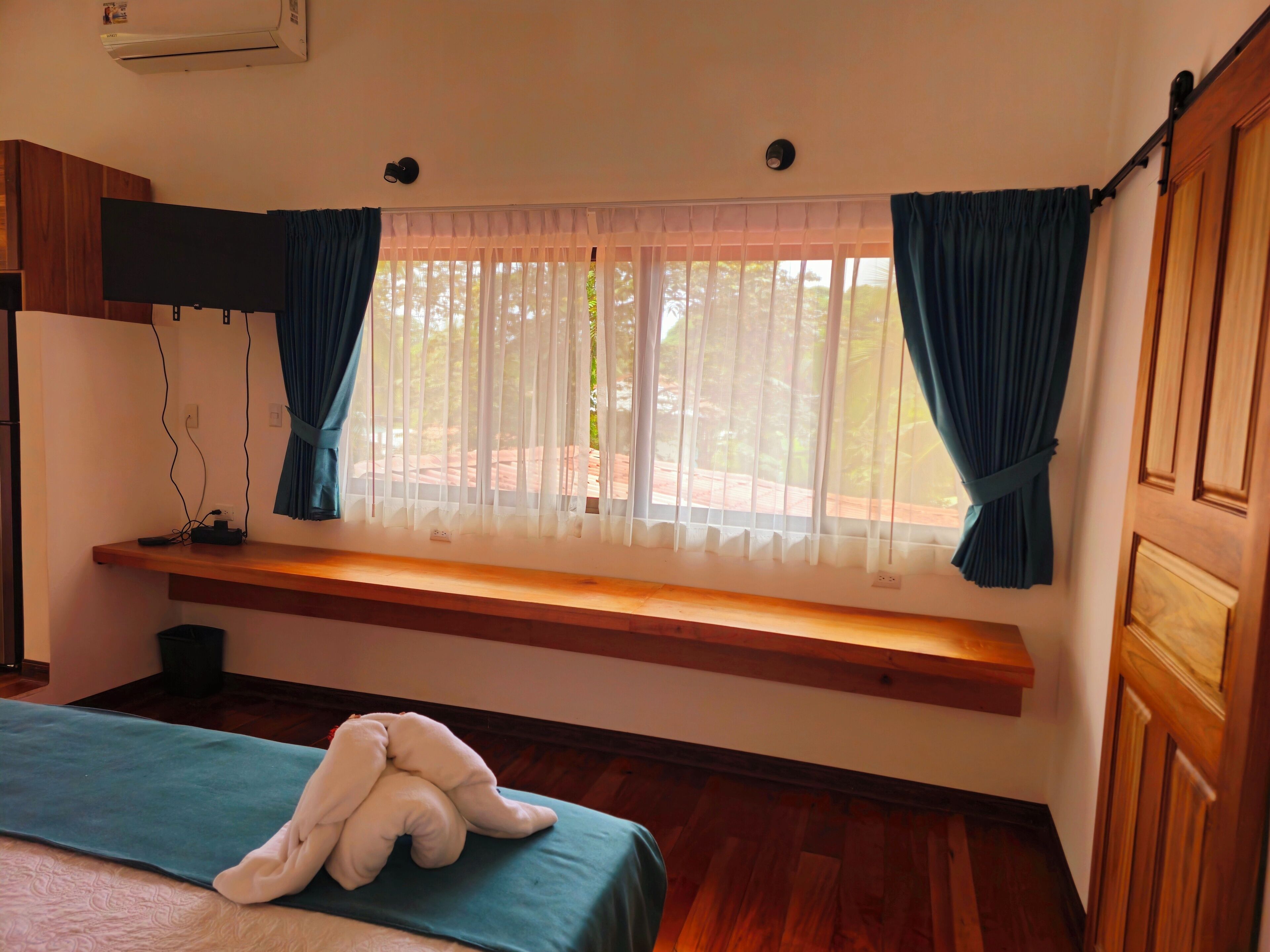 Une chambre avec un banc en bois près de la fenêtre, un lit recouvert d’une couverture bleue et un ours en peluche blanc posé sur le lit.