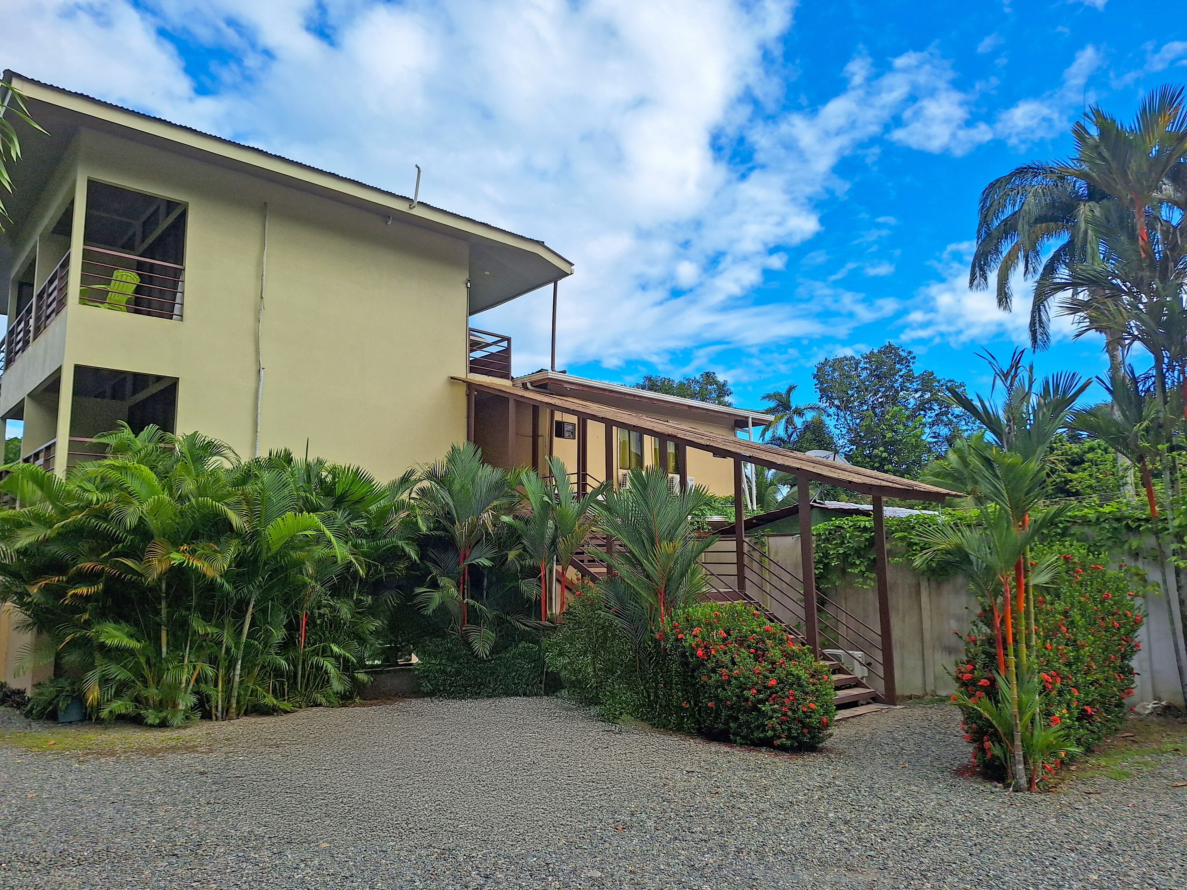 A two-story building with a balcony, surrounded by lush greenery and a gravel driveway.