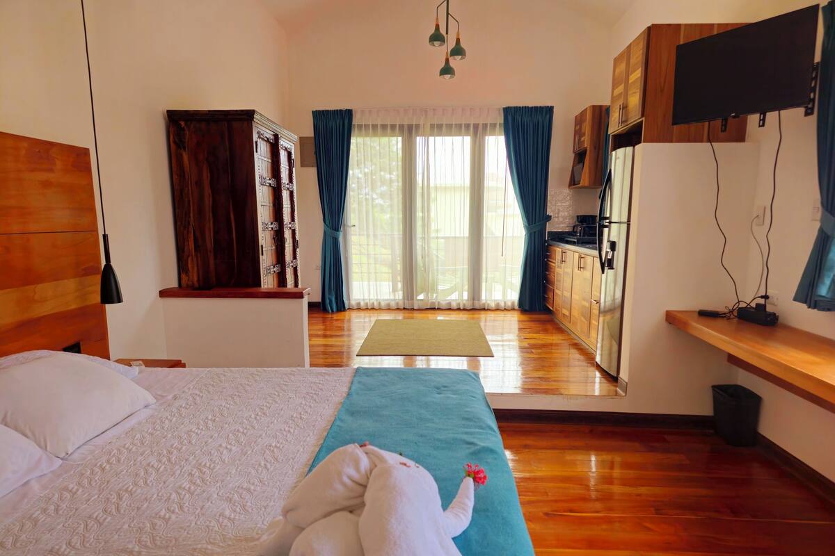 Une chambre d’hôtel avec un lit, une tête de lit en bois, une kitchenette et une vue sur l’extérieur.