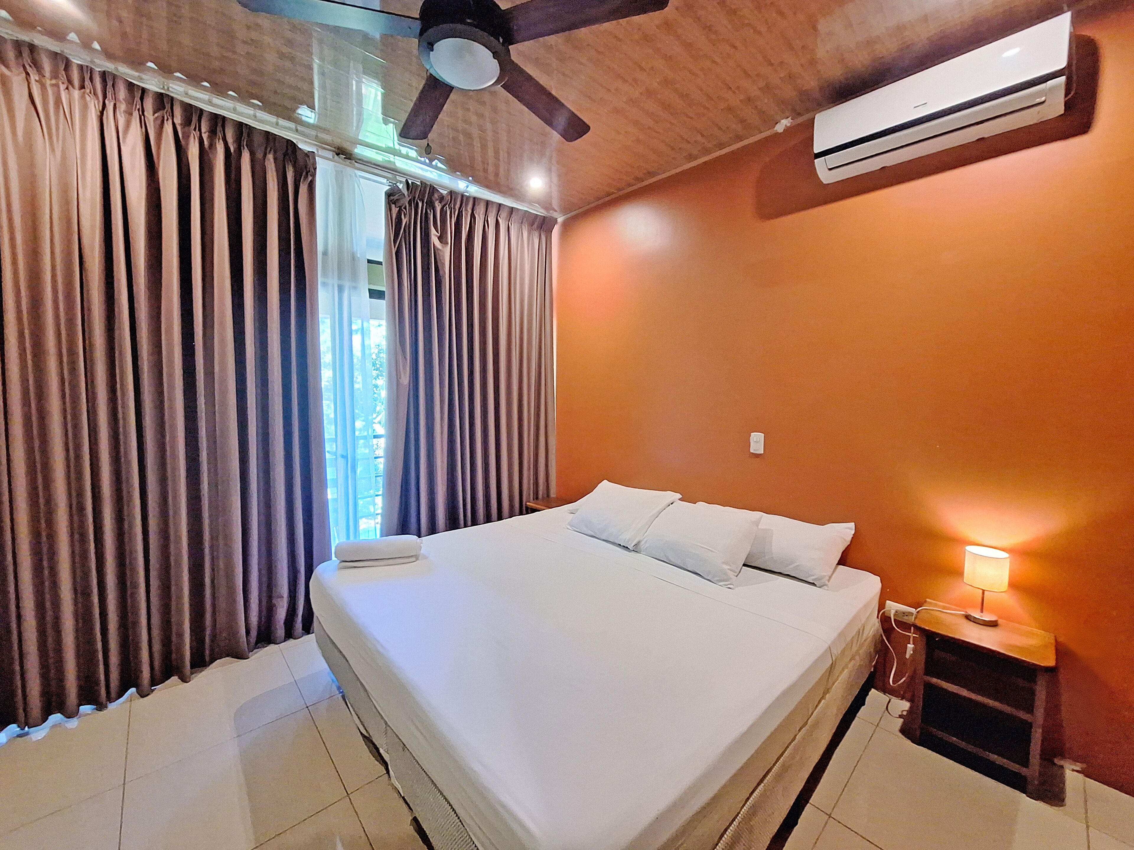 Un dormitorio con cama, cortinas, ventilador de techo, aire acondicionado y lámpara de noche.