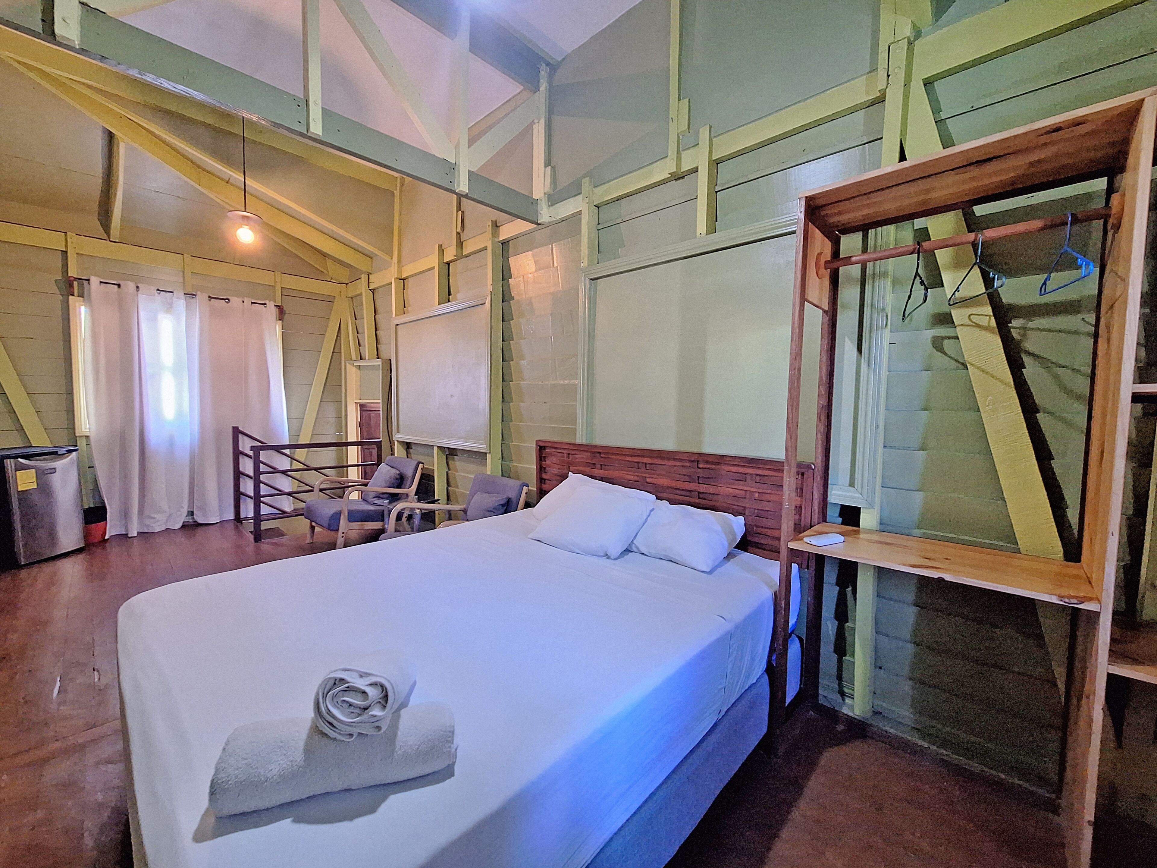 Una habitación con una cama grande, un cabecero de madera, un escritorio con una silla, una ventana con cortinas y una lámpara colgante.