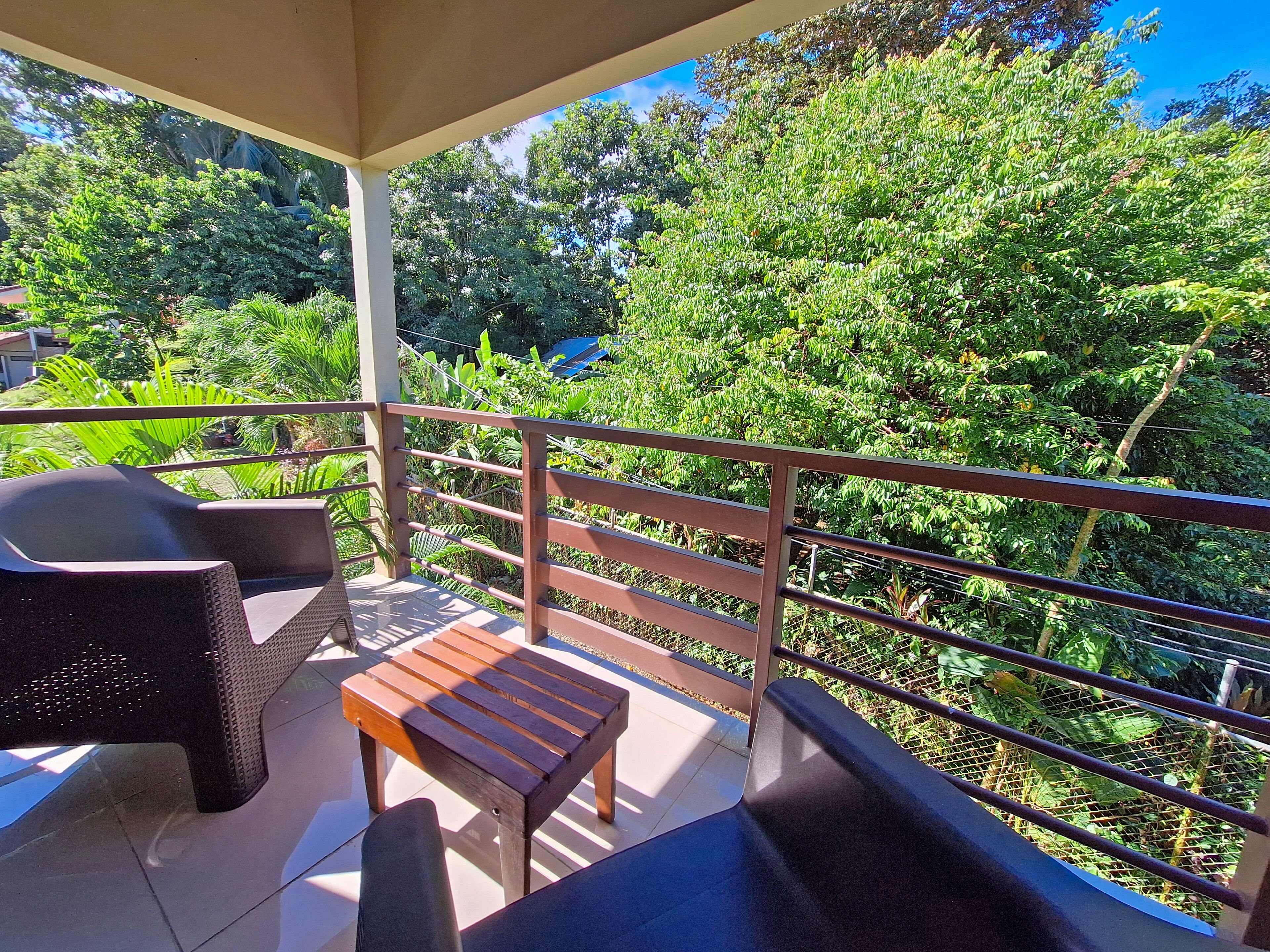 A balcony with a wooden table and two chairs, surrounded by lush greenery and a metal railing.