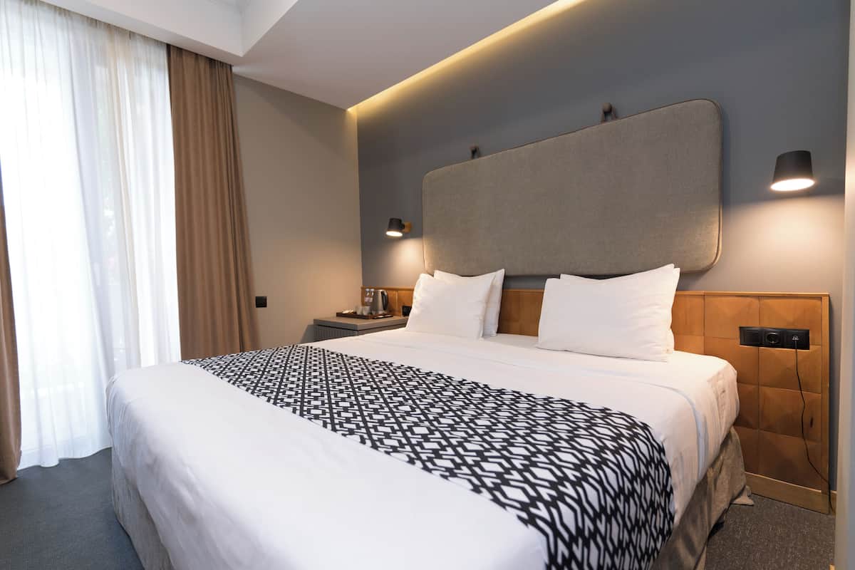 A modern hotel room with a large bed, a headboard with integrated lighting, a bedside table with a lamp, and a view of the outdoors through sheer curtains.